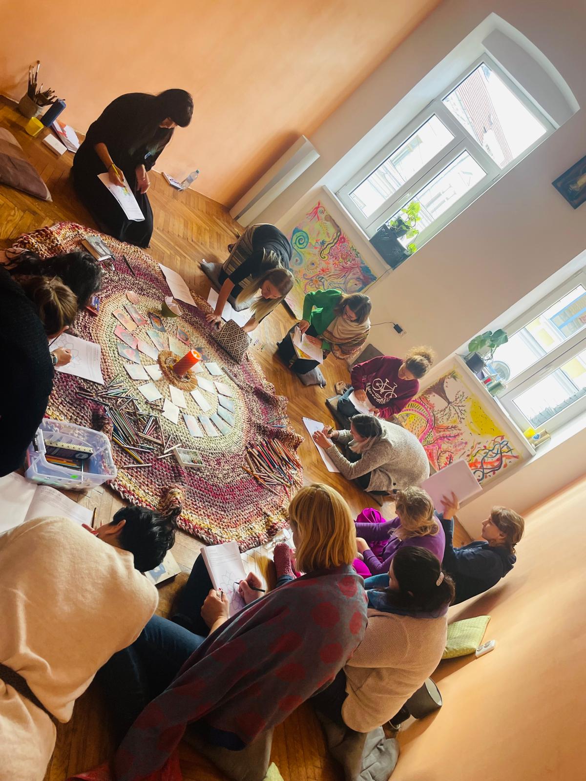 Cerc de participante așezate pe podea, în jurul unui covor cu cărți și creioane colorate