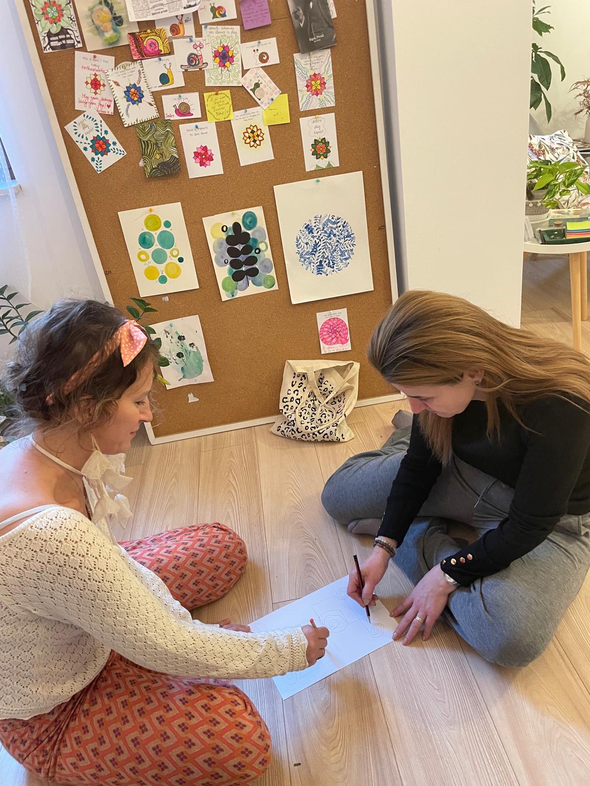 Două participante desenând împreună pe podea, în fața unui panou cu lucrări MindfulArt®